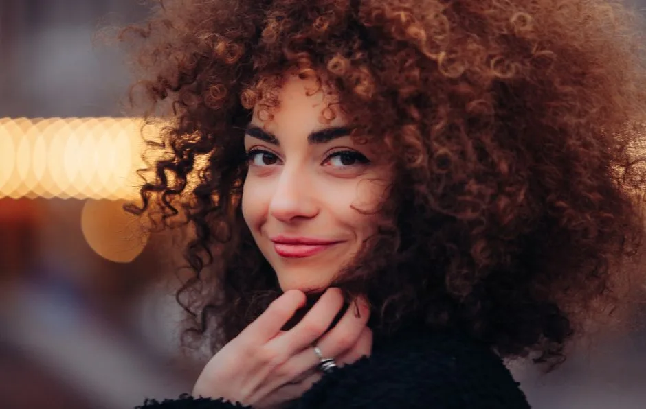 Close-up portrait of a woman with curly hair smiling warmly in soft, natural light.