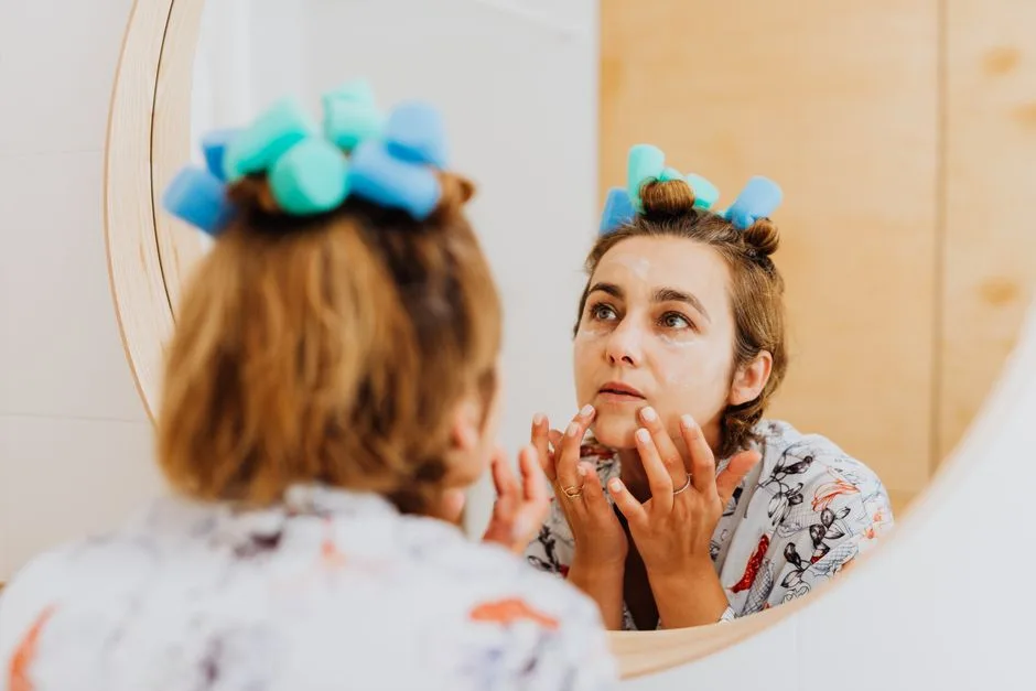 Woman with curlers applying cream in front of a mirror indoors.