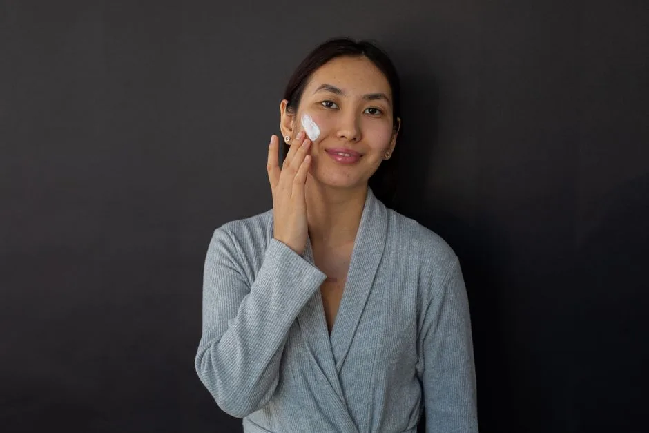 Positive ethnic female in bathrobe applying moisturizing cream on cheek during daily beauty routine against black background and looking at camera
