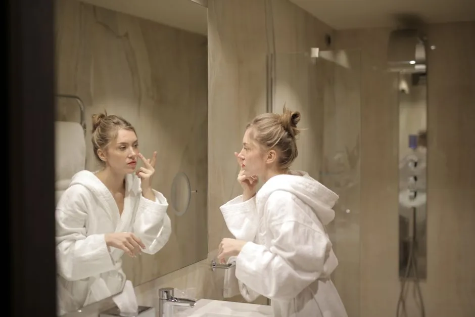 A woman in a white bathrobe applies moisturizer while looking at her reflection in the bathroom mirror.